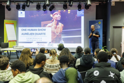Aurelien Duarte sur la scène du Virtuoz Club de Villeneuve-la-Garenne tient dans sa main un micro pour présenter sa conférence. - Agrandir l'image 6 sur 6, fenêtre modale