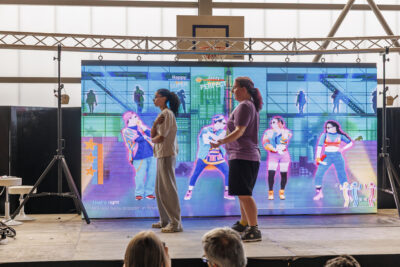 Deux jeunes sont sur une scène et jouent à Just Dance dans le cadre du salon de jeux vidéos de Villeneuve-la-Garenne - Agrandir l'image 5 sur 6, fenêtre modale