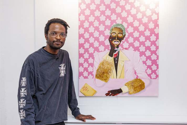 L'artiste Jules Be Kuti pose à côté d'un de ses tableaux qui représente un homme noir sur une toile rose.