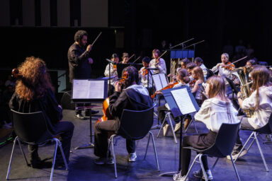 Un maître d'orchestre guide plusieurs jeunes sont sur scène en train de jouer d'un instrument dans le cadre du gala de fin d'année de l'école de musique Claude-Debussy à Villeneuve-la-Garenne.