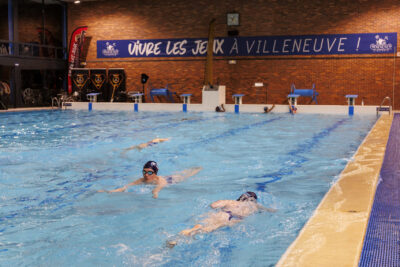 Une piscine intérieure avec des nageurs et un mur portant une bannière "Vivre les Jeux à Villeneuve !". - Agrandir l'image 4 sur 7, fenêtre modale