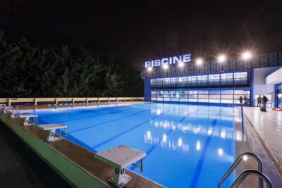 Une piscine extérieure illuminée la nuit avec un bâtiment vitré et le mot "Piscine" en lettres lumineuses. - Agrandir l'image 5 sur 7, fenêtre modale