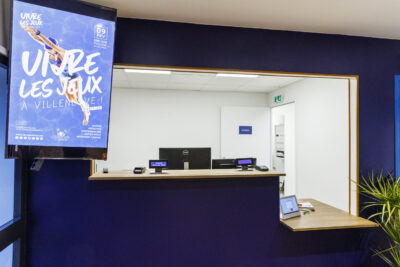 Un espace d'accueil moderne avec un guichet et un écran annonçant un l'événement "Vivre les jeux à Viulleneuve". - Agrandir l'image 7 sur 7, fenêtre modale