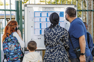 Un homme, une femme et deux enfants regardent un tableau des classes devant une école - Agrandir l'image 2 sur 6, fenêtre modale