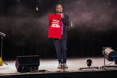L'humoriste Kosh sir scène tenant son tee shirt avec écrit faut pas louper l'Kosh - Agrandir l'image 3 sur 6, fenêtre modale