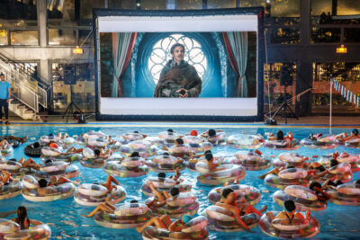 Plusieurs personnes sont assis dans une bouée gonflable à la piscine municipale de Villeneuve-la-Garenne face à un écran géant pour regarder un film à l'événement ciné piscine - Agrandir l'image 4 sur 6, fenêtre modale